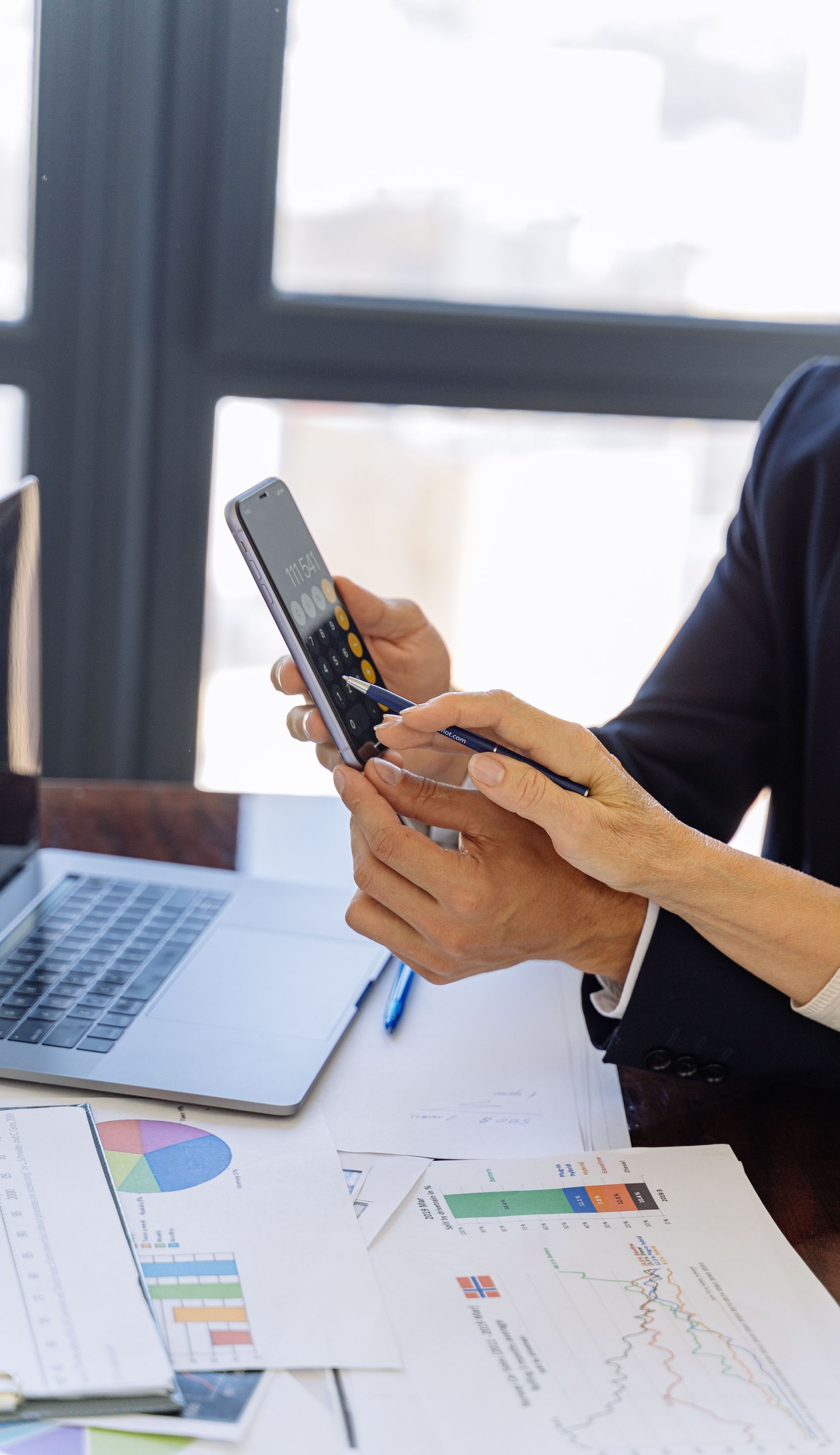 Business professionals reviewing financial reports on a smartphone at a desk with papers and a laptop in an office setting.